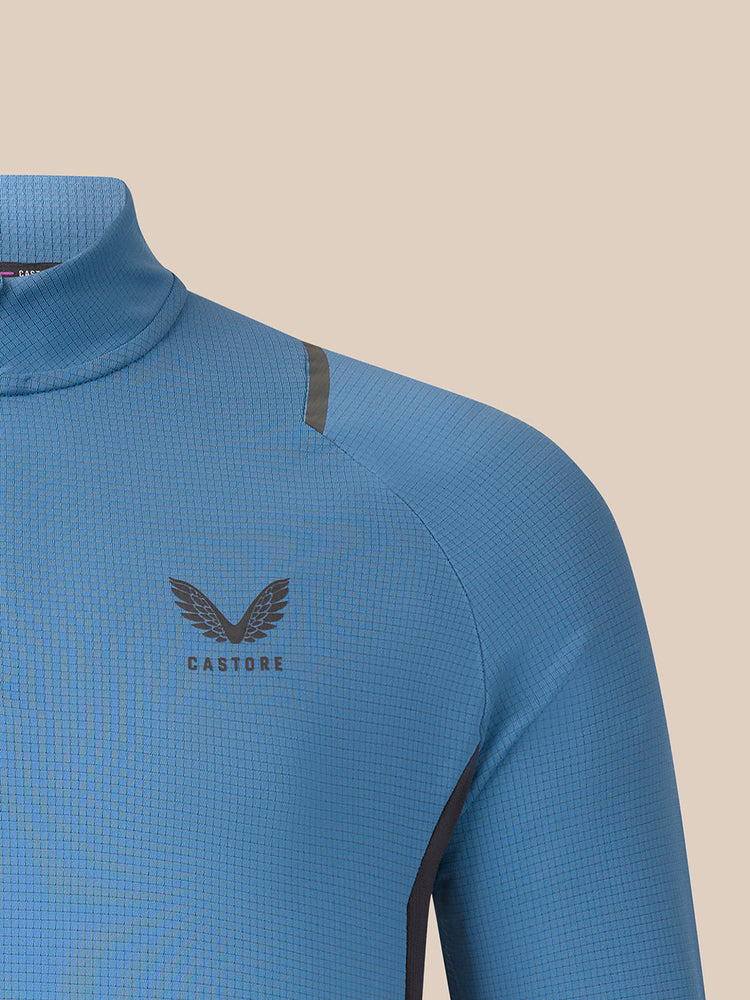 Close-up of a light blue long-sleeve athletic top with a dark gray wing logo and the word CASTORE on the chest, set against a solid beige background.