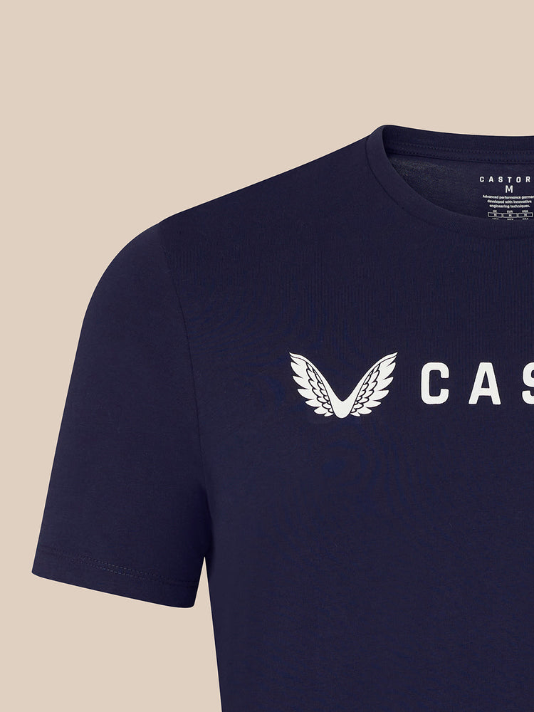 Close-up of a navy blue short-sleeve t-shirt with a white winged logo and partial 'CAS' text on the chest, shown against a light beige background.