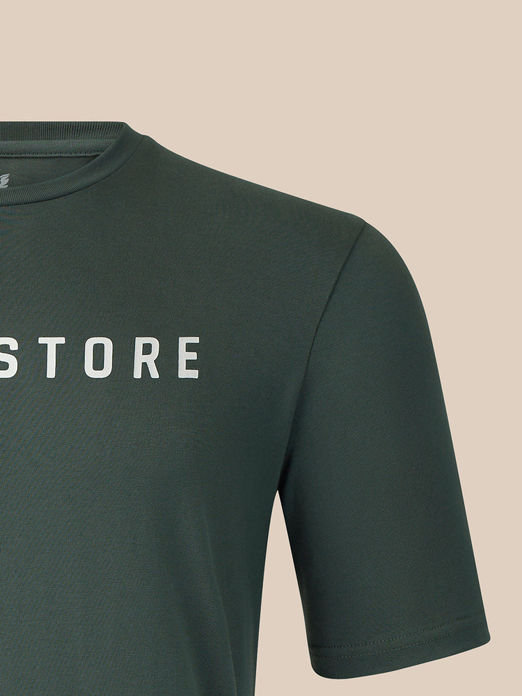 Close-up of a dark green short-sleeve t-shirt with the word 'CASTORE' printed in white capital letters across the chest, set against a solid beige background.